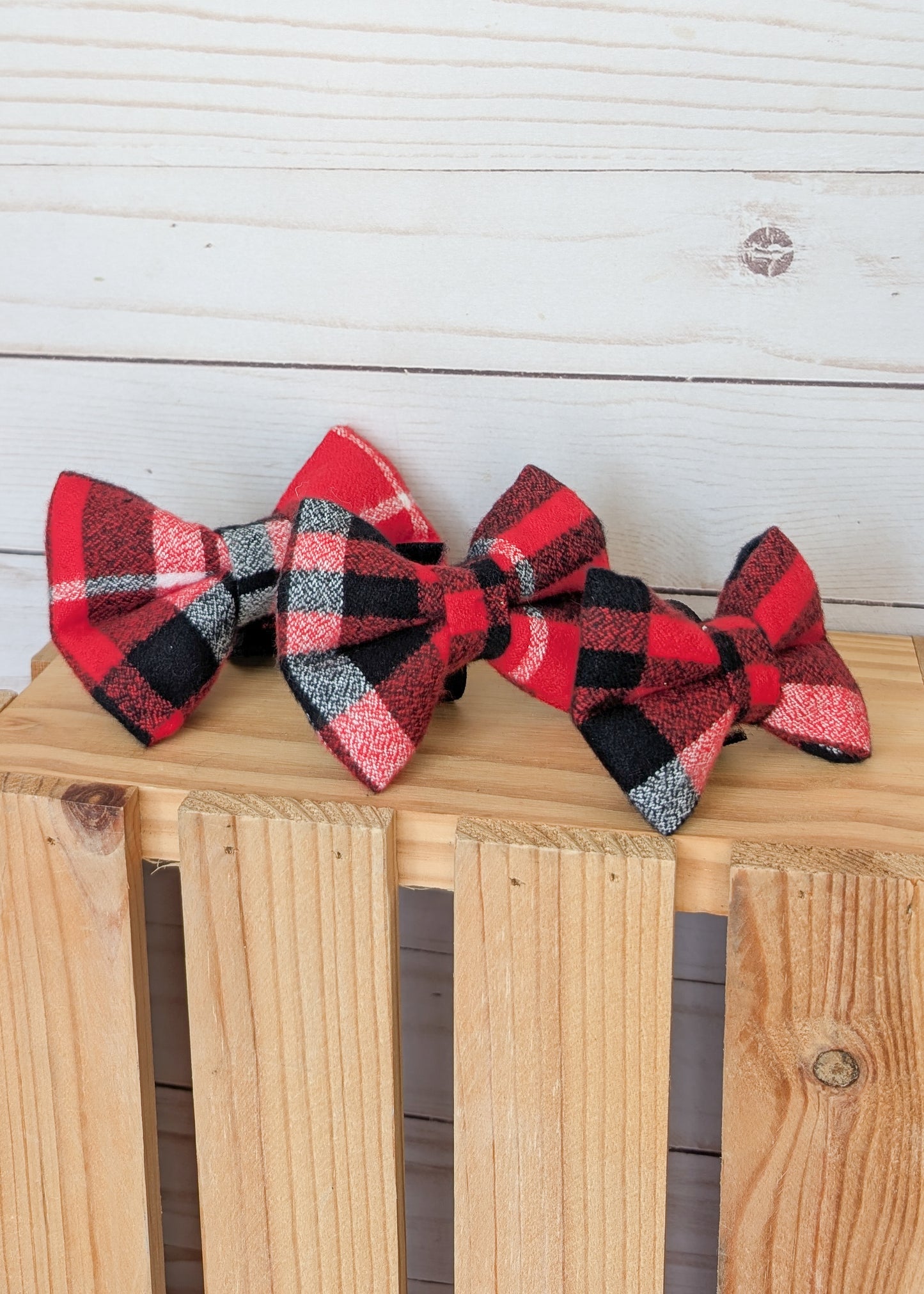 Three red, black, and white plaid bow ties arranged on a wooden crate against a white wood plank background.