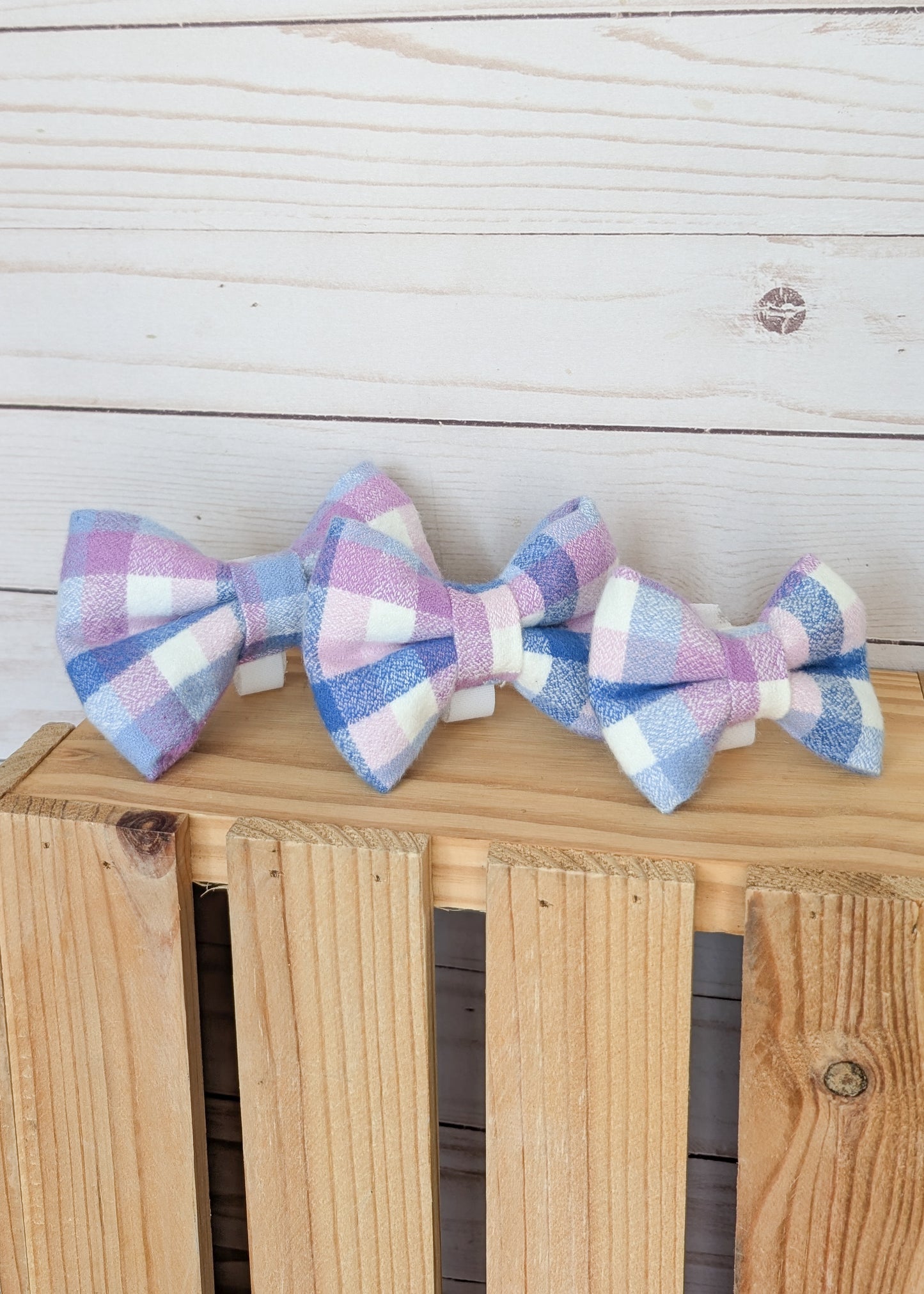 Three fabric bow ties with a pastel gingham plaid pattern in pink, purple, blue, and white, displayed on a wooden crate against a white wood plank background.