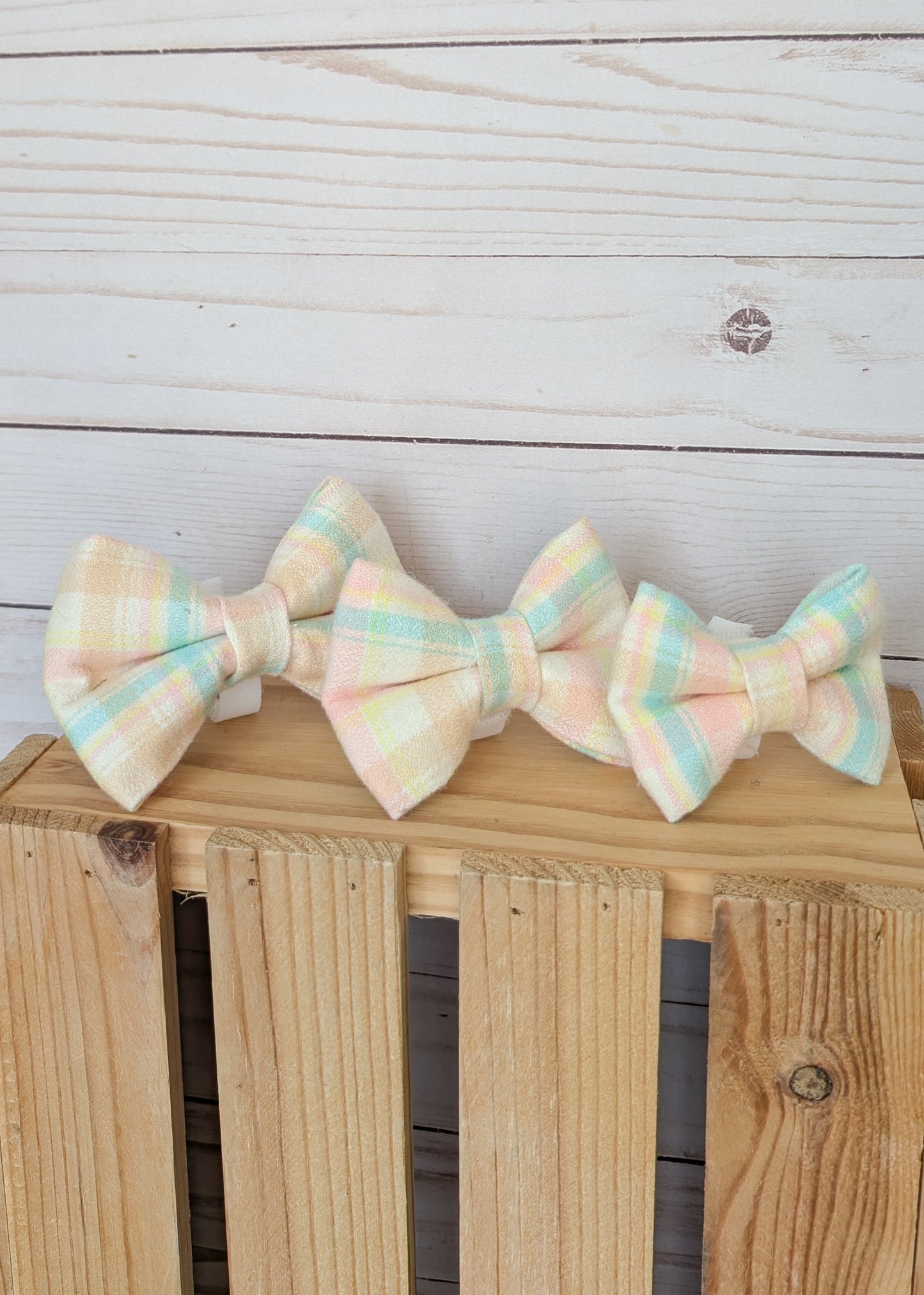 Three pastel plaid bow ties in soft peach, mint green, and cream are arranged on a wooden crate against a whitewashed wood plank background.
