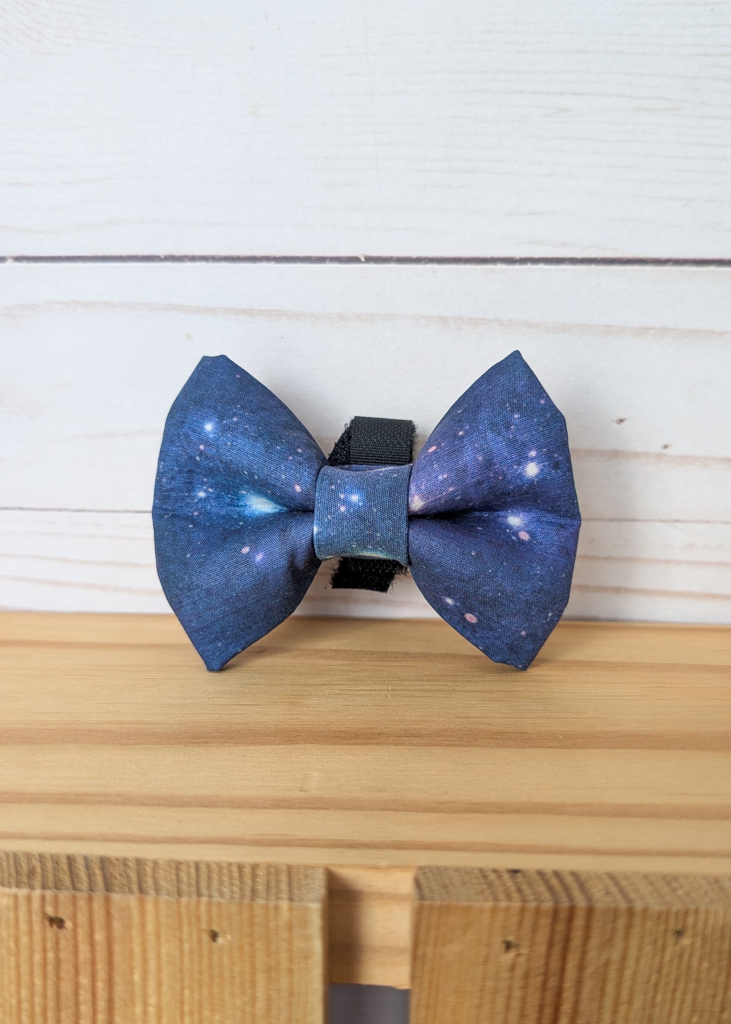 A galaxy-printed bow tie with a blue and purple cosmic pattern, resting on a light wooden surface against a white wood plank background.
