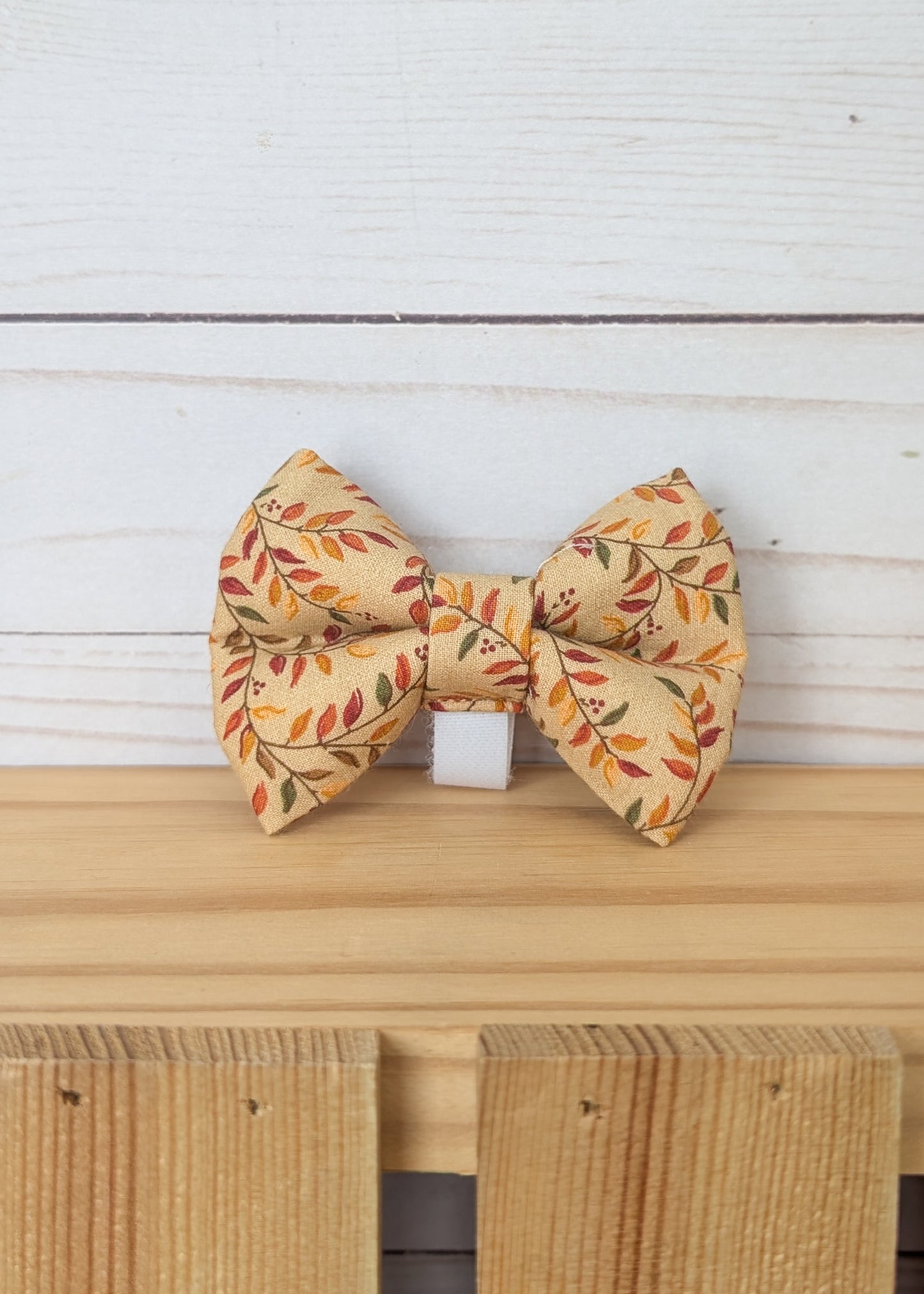A small fabric bow tie with an autumn leaf pattern in orange, red, and green on a warm beige background, displayed on a wooden surface against a white wood plank backdrop.