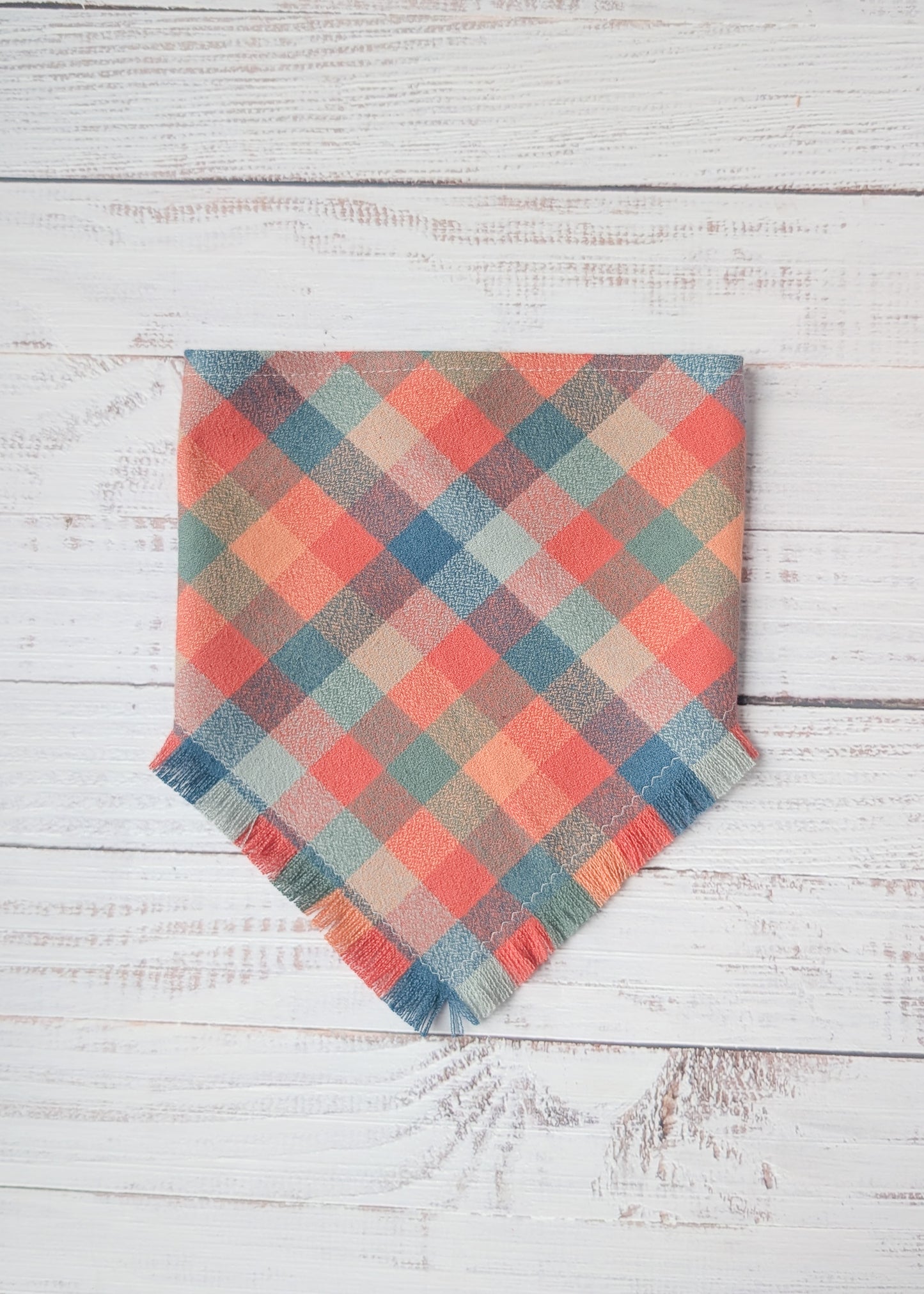 Plaid flannel bandana with frayed edges with dusty pink, light blue, orange colors,  on a wooden surface