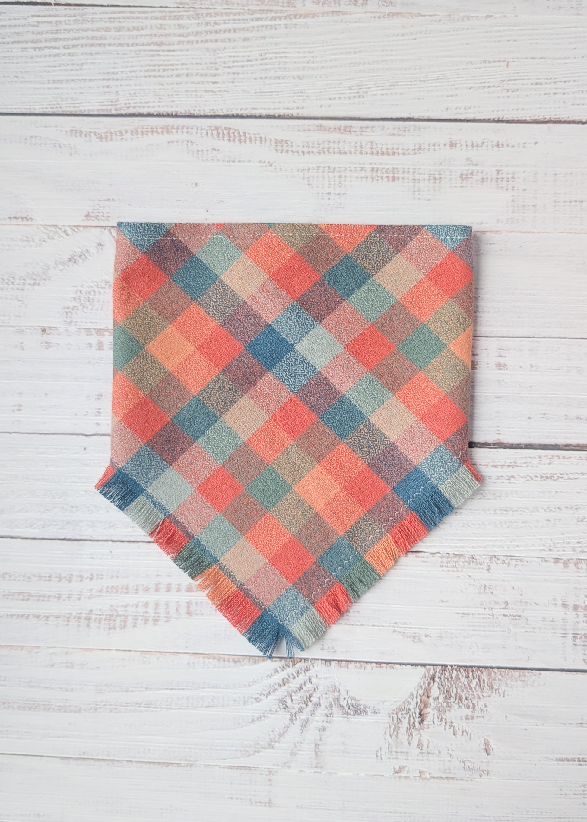 Plaid flannel bandana with frayed edges with dusty pink, light blue, orange colors,  on a wooden surface