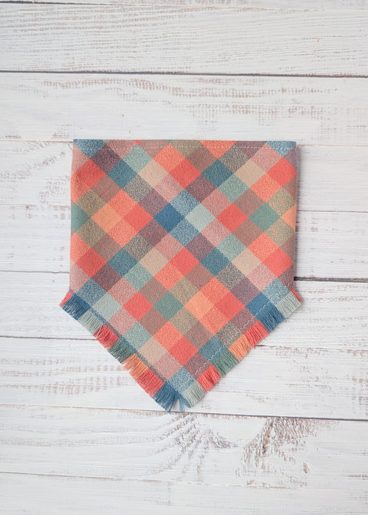 Plaid flannel bandana with frayed edges with dusty pink, light blue, orange colors,  on a wooden surface