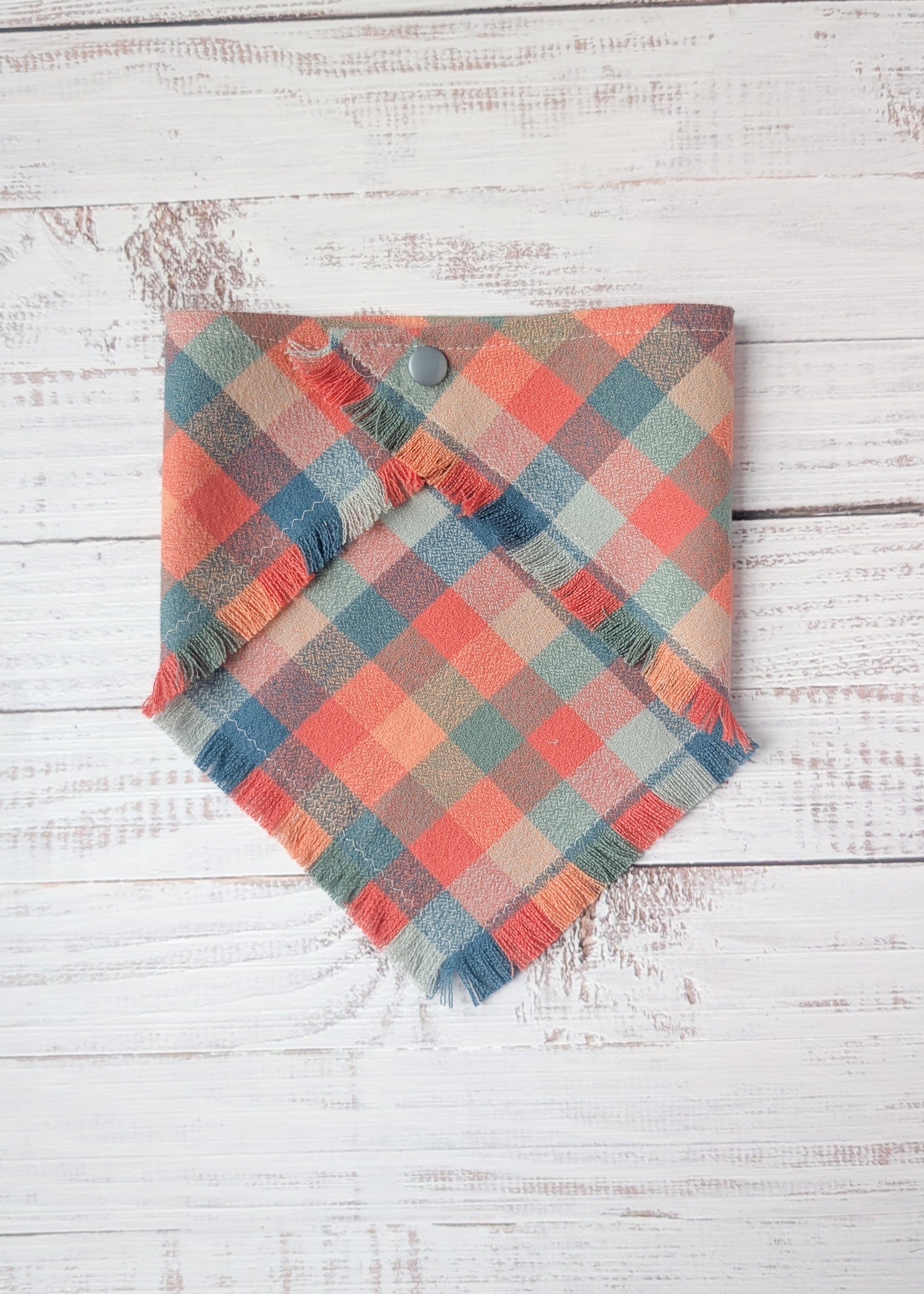 Plaid flannel bandana with frayed edges with dusty pink, light blue, orange colors,  on a wooden surface