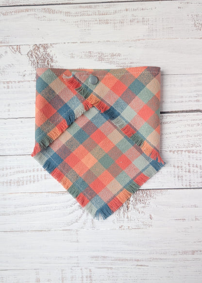 Plaid flannel bandana with frayed edges with dusty pink, light blue, orange colors,  on a wooden surface