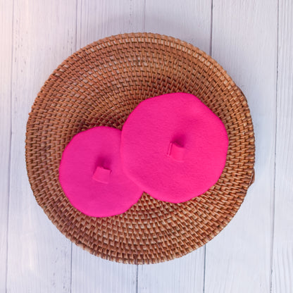 Two pink dog berets, shown from the top, on a wicker circle on a white wood background