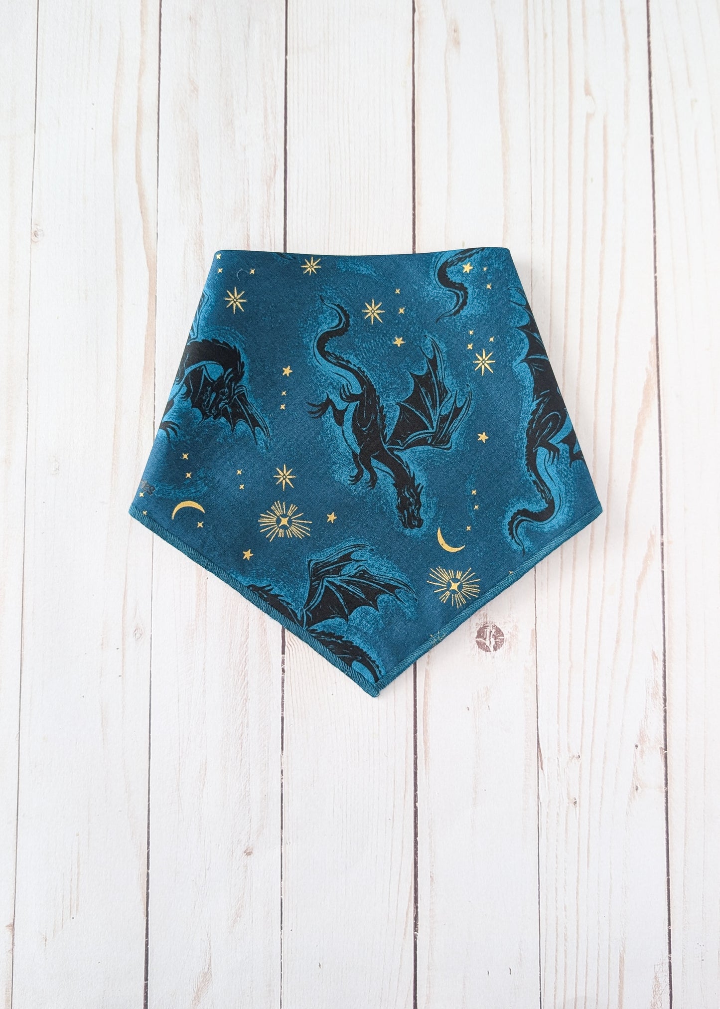 A triangular bandana-style pet bib or scarf in teal blue, featuring a printed pattern of black dragons, gold stars, and gold crescent moons, laid flat on a white wooden plank background.