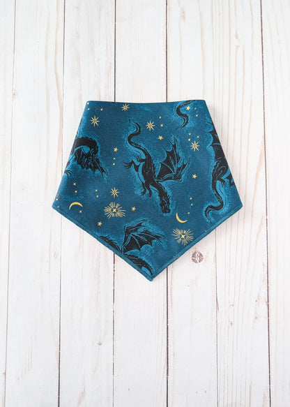A triangular bandana-style pet bib or scarf in teal blue, featuring a printed pattern of black dragons, gold stars, and gold crescent moons, laid flat on a white wooden plank background.