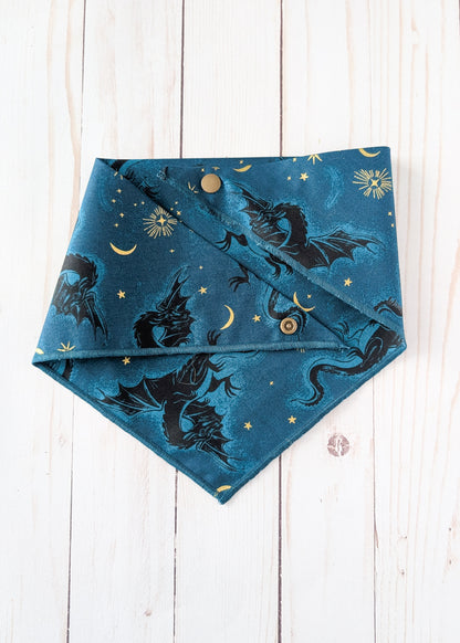A triangular bandana-style pet collar or bib in teal blue fabric with a printed pattern of black dragons, gold stars, and crescent moons, laid flat on a white wood plank background.