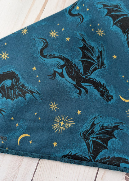 A close-up of a blue fabric with a printed pattern of black dragons, gold stars, and gold crescent moons, laid flat on a light wood surface.