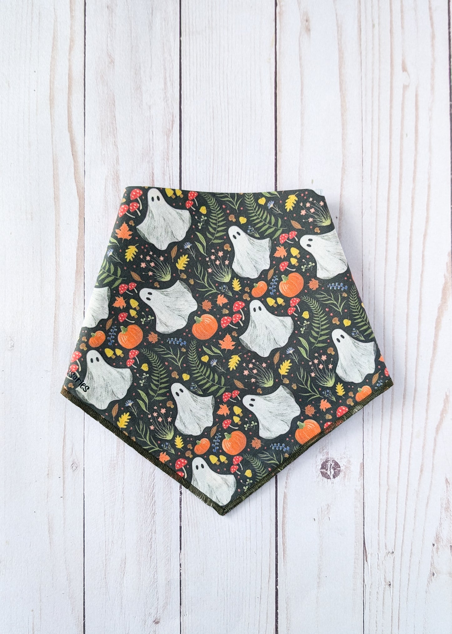 A triangle bandana with a Halloween pattern of white ghosts, orange pumpkins, and green foliage on a dark background, laid flat on a white wood plank surface.