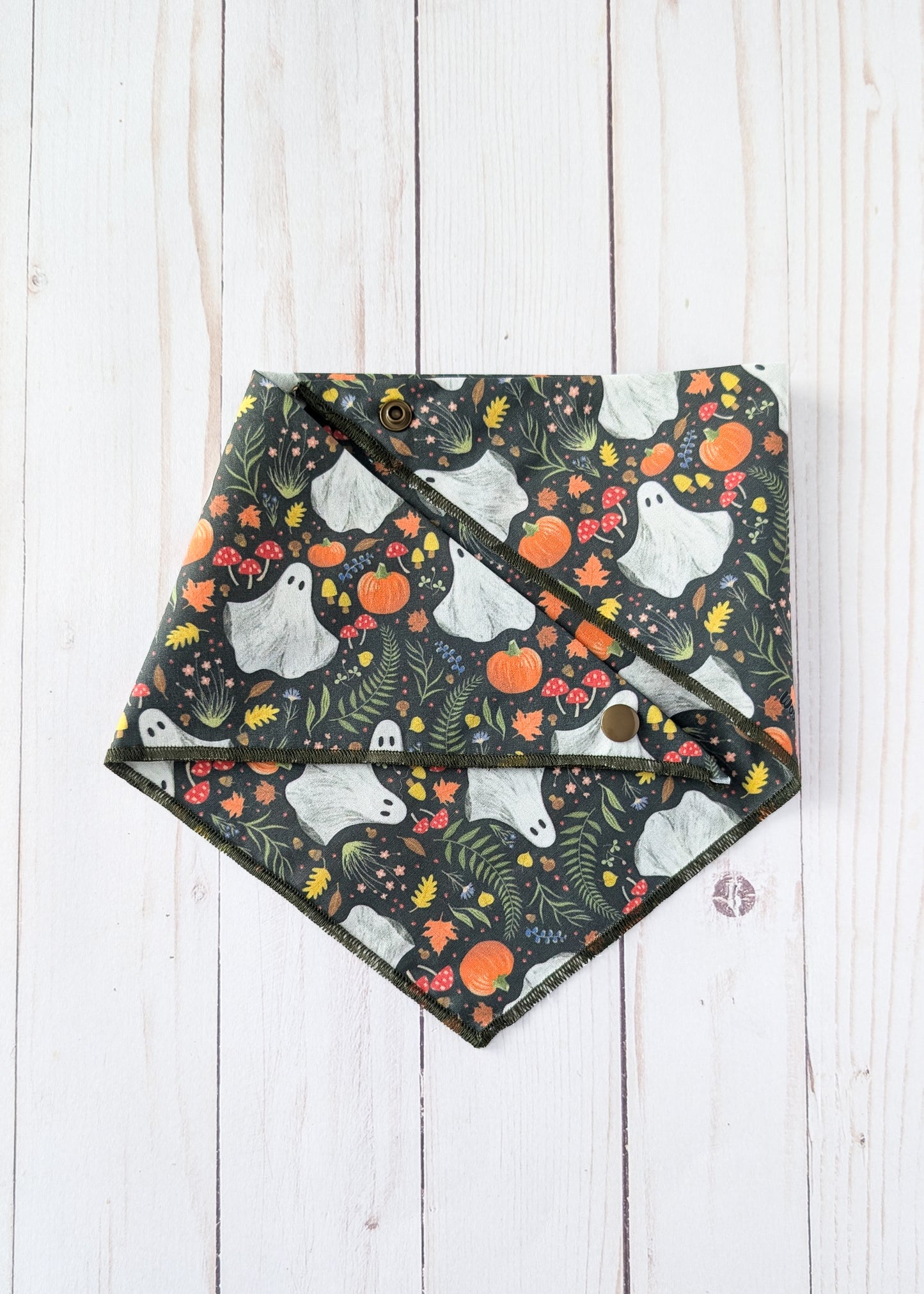 A triangle bandana with a Halloween pattern of white ghosts, orange pumpkins, and green foliage on a dark background, laid flat on a white wood plank surface.