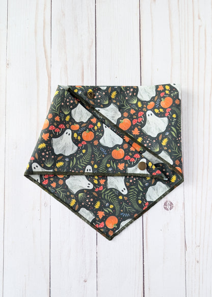 A triangle bandana with a Halloween pattern of white ghosts, orange pumpkins, and green foliage on a dark background, laid flat on a white wood plank surface.