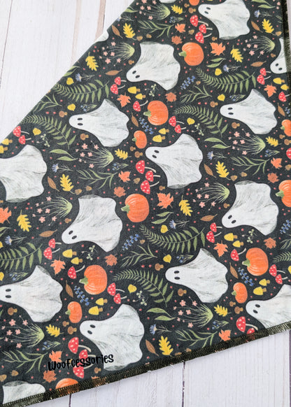 A close-up of a fabric or textile with an all-over Halloween pattern featuring white ghosts, orange pumpkins, green leaves, and small red and yellow floral details on a dark background, laid diagonally on a white wood surface.