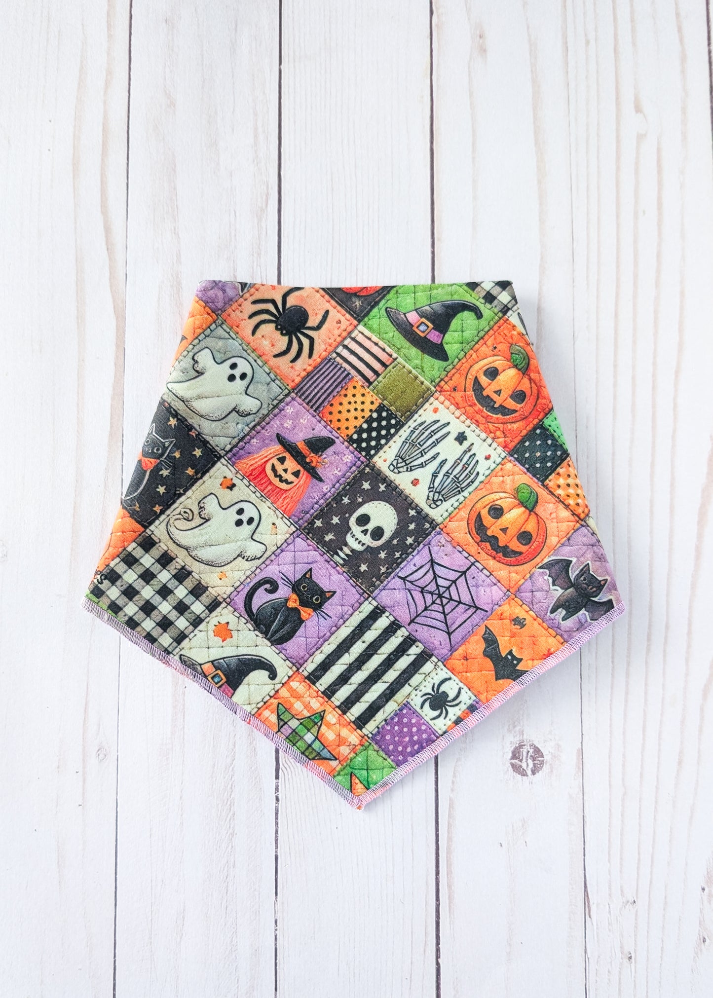 A triangular bandana with a colorful Halloween patchwork print, displayed flat on a white wood plank background.