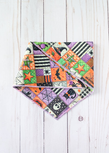 A triangular bandana with a colorful Halloween patchwork print, displayed flat on a white wood plank background.