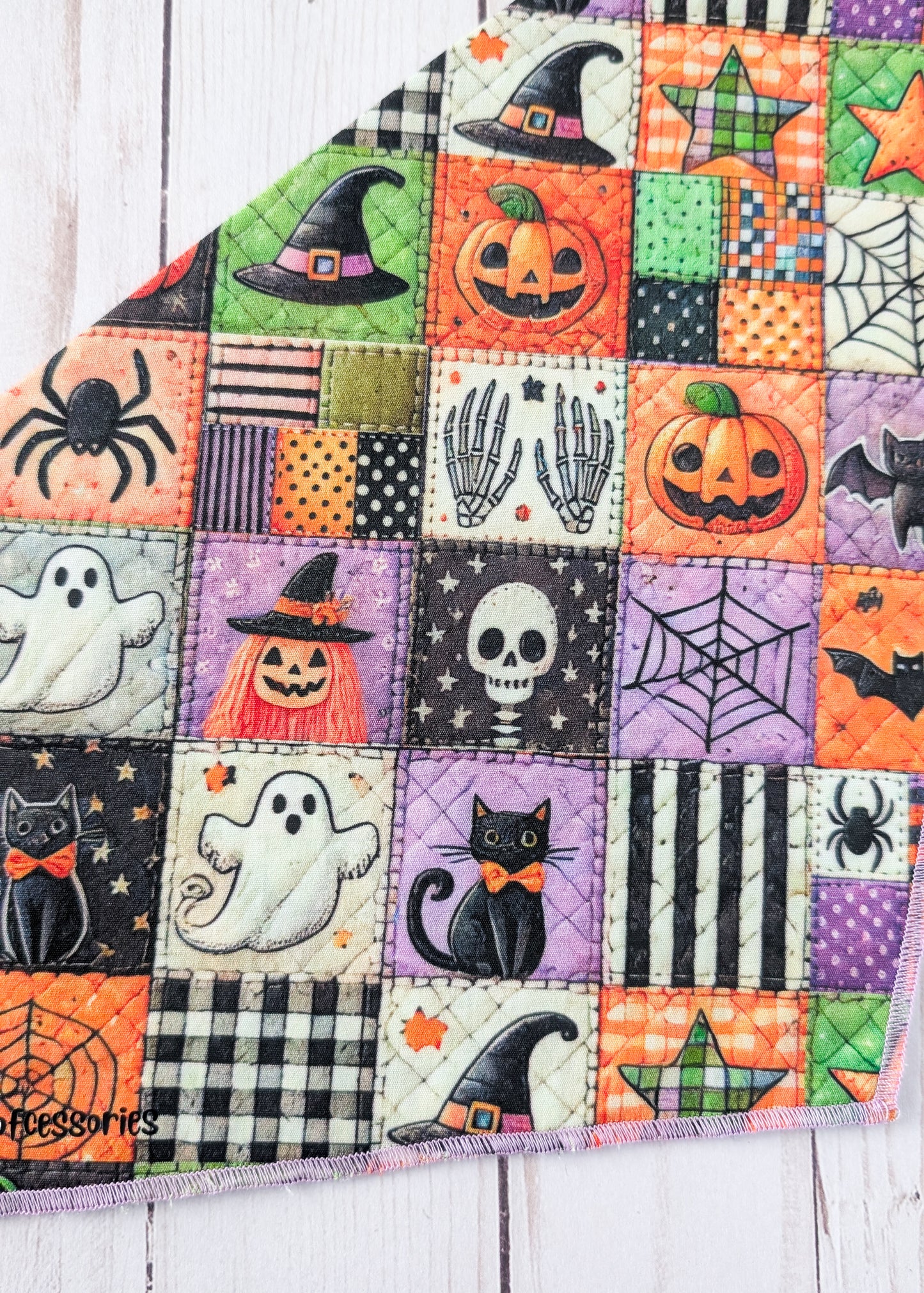 A close up of a triangular bandana with a colorful Halloween patchwork print, displayed flat on a white wood plank background.