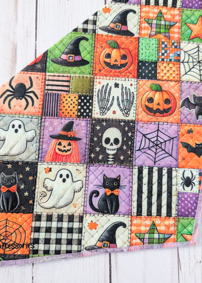 A close up of a triangular bandana with a colorful Halloween patchwork print, displayed flat on a white wood plank background.