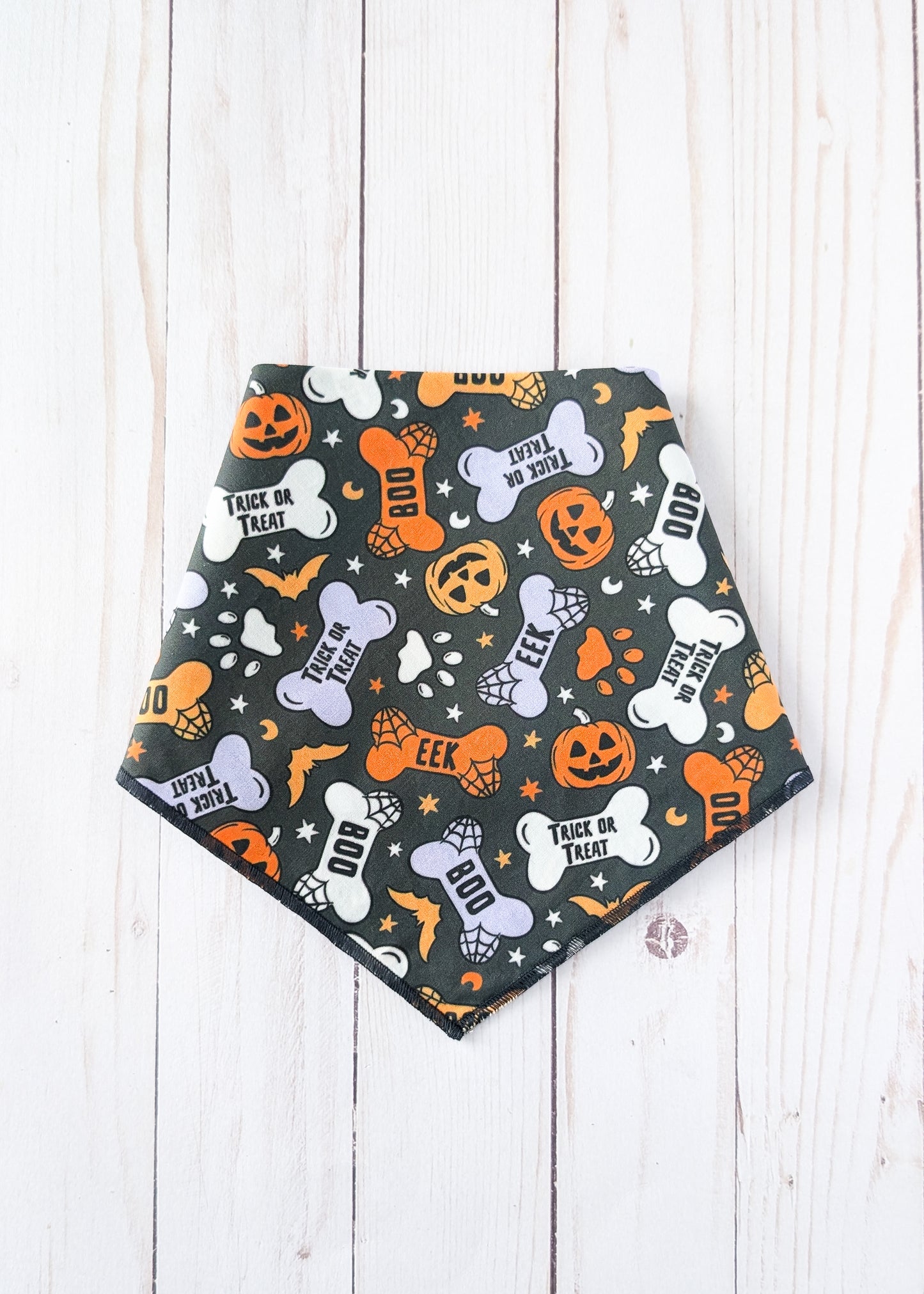 A triangular dog bandana with a Halloween print, laid flat on a white wood plank background. The bandana is black with orange, white, and light purple patterns of pumpkins, bones, ghosts, and the text 'TRICK OR TREAT'.
