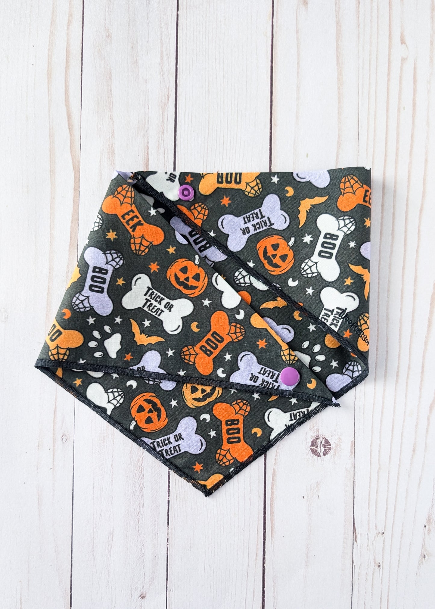 A triangular pet bandana with a black background and a colorful Halloween pattern featuring orange pumpkins, white bones, purple tombstones, and text like 'BOO' and 'TRICK or TREAT', laid flat on a white wood plank background.