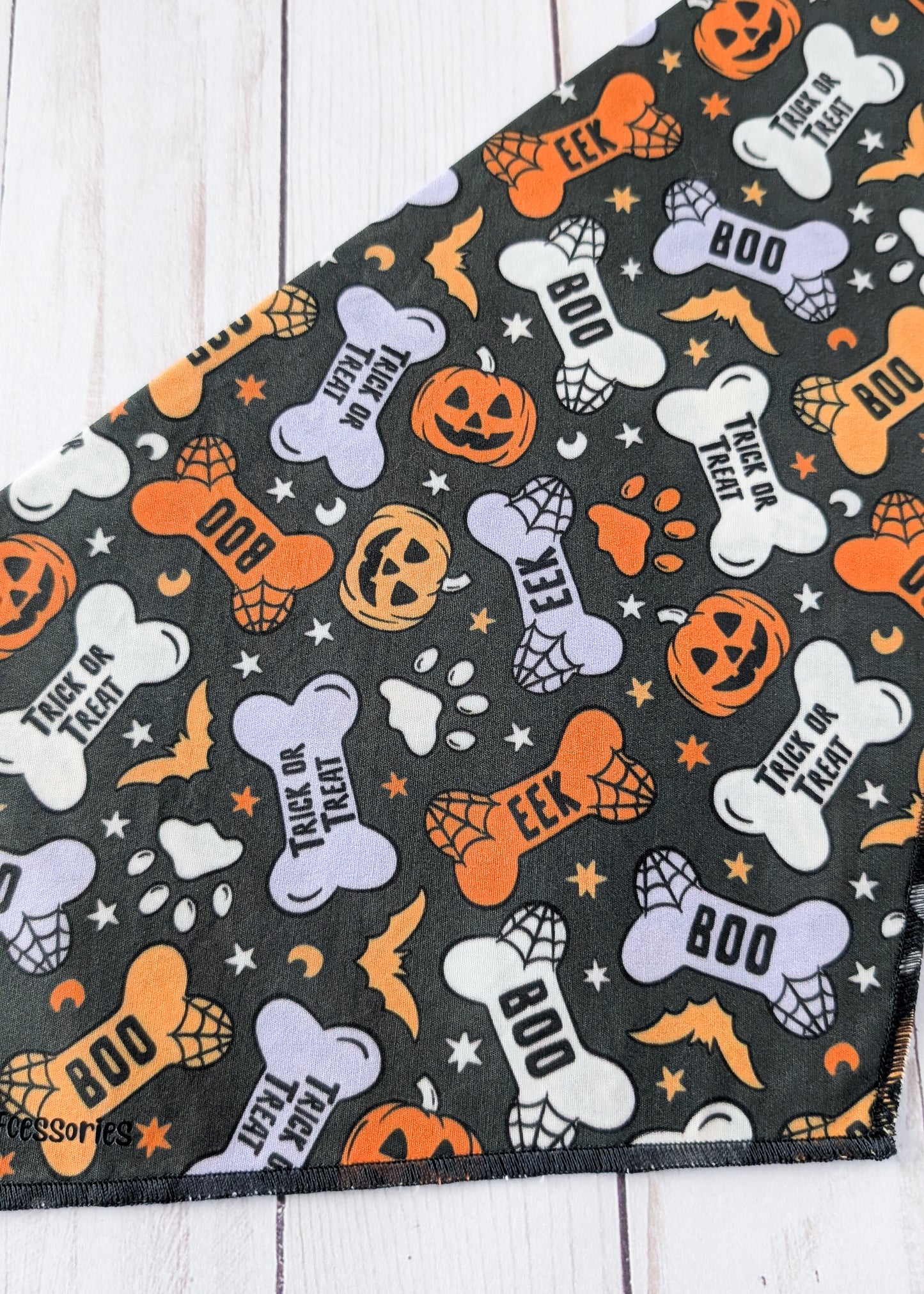 A close-up of a black fabric bandana or scarf featuring a Halloween pattern with orange pumpkins, white bones, bats, spiderwebs, and text like 'BOO', 'TRICK OR TREAT', and 'EEK'.