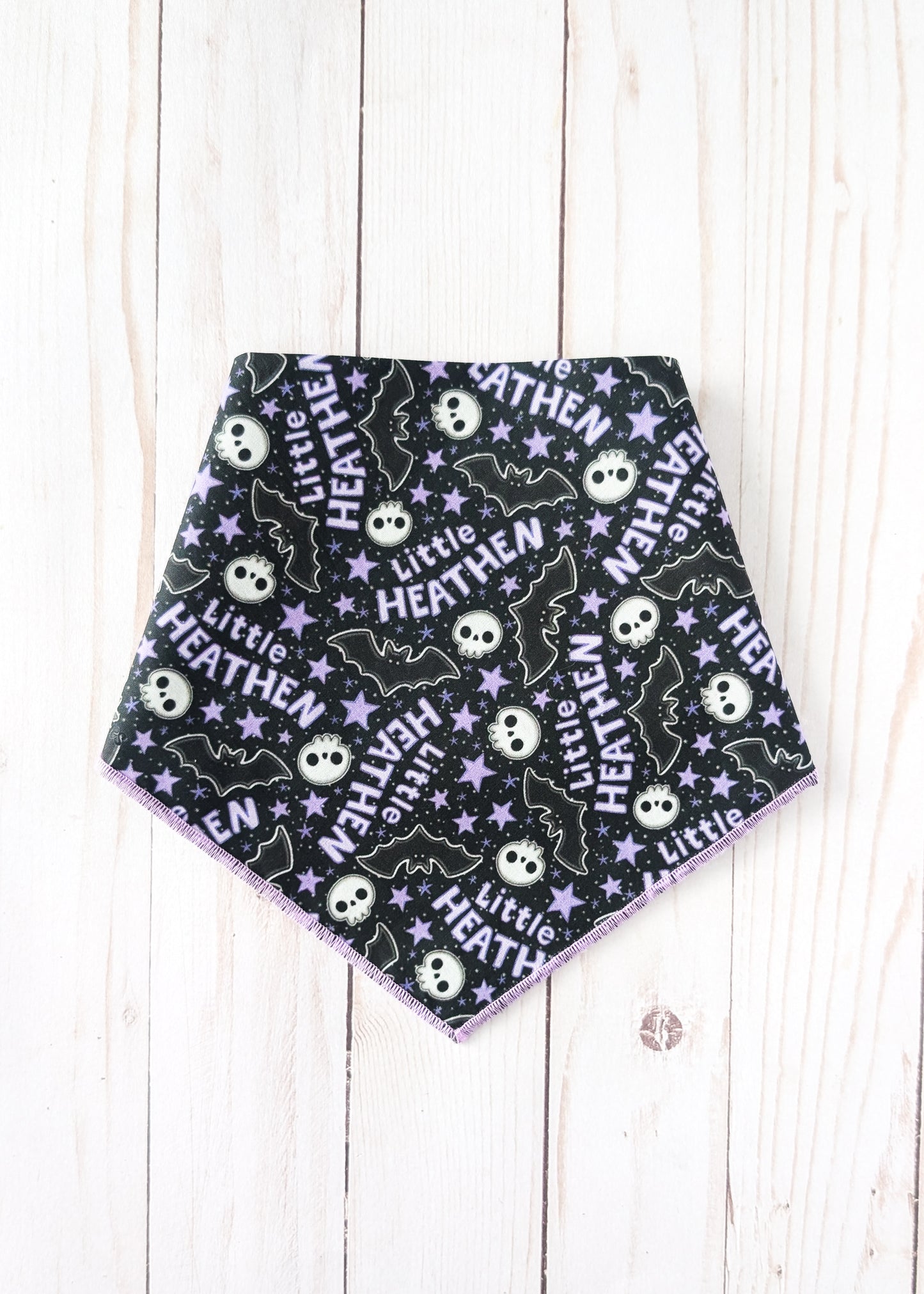 A triangular pet bandana with a black background featuring a repeating Halloween pattern of white skulls, purple bats, and white stars with the text 'Little HEATHEN'. The bandana is laid flat on a light-colored wooden plank background.