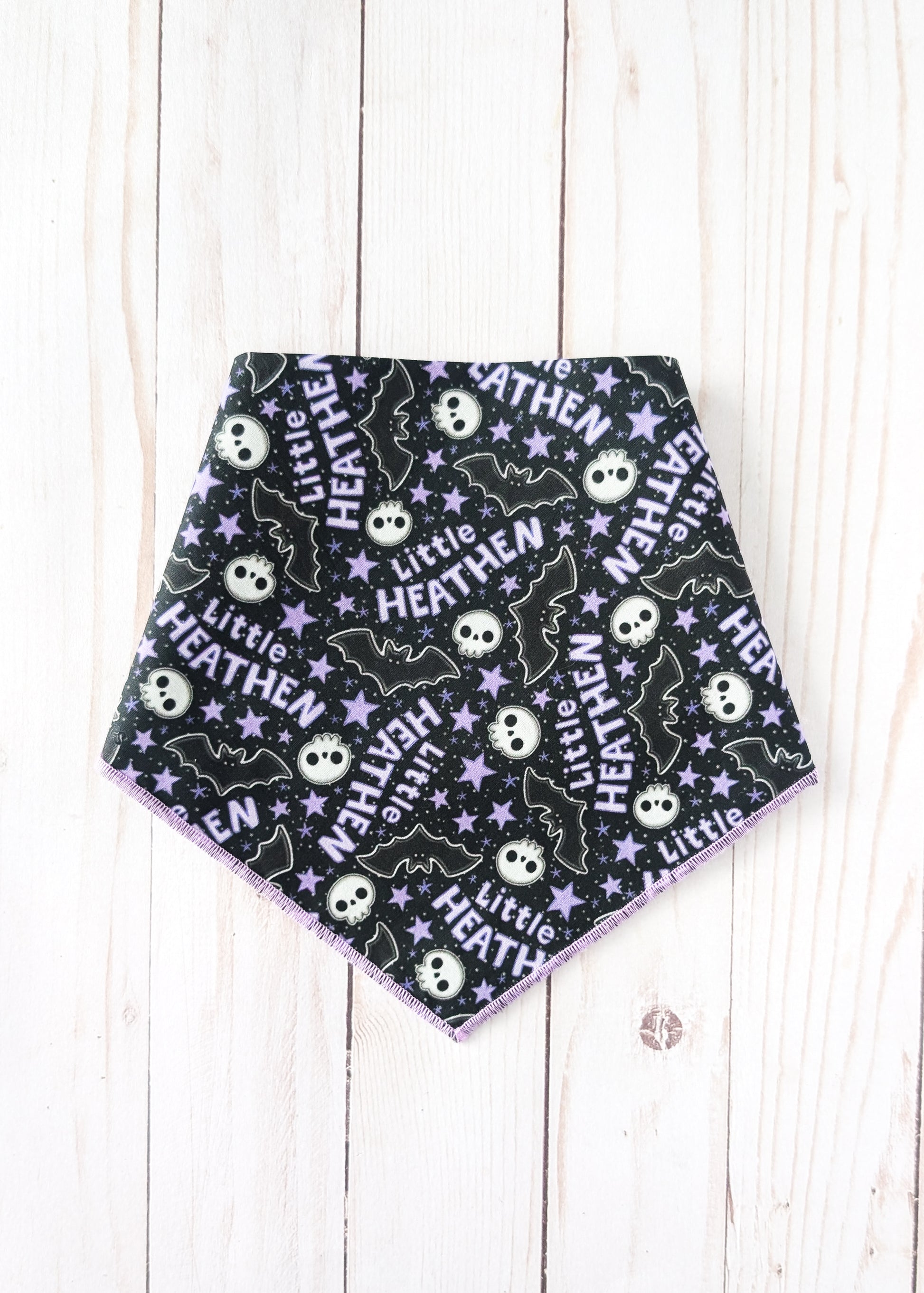 A triangular pet bandana with a black background featuring a repeating Halloween pattern of white skulls, purple bats, and white stars with the text 'Little HEATHEN'. The bandana is laid flat on a light-colored wooden plank background.