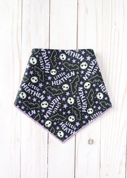 A triangular pet bandana with a black background featuring a repeating Halloween pattern of white skulls, purple bats, and white stars with the text 'Little HEATHEN'. The bandana is laid flat on a light-colored wooden plank background.