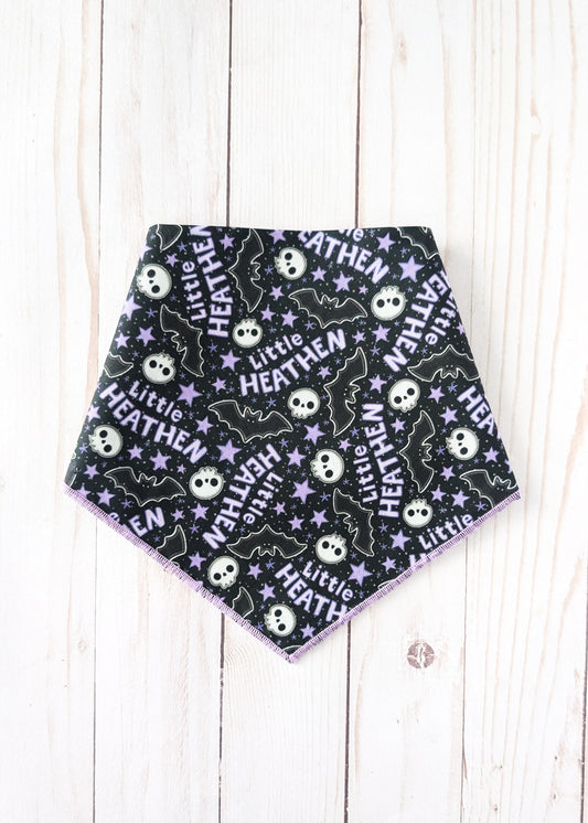 A triangular pet bandana with a black background featuring a repeating Halloween pattern of white skulls, purple bats, and white stars with the text 'Little HEATHEN'. The bandana is laid flat on a light-colored wooden plank background.