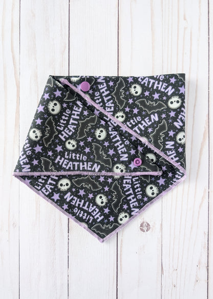 A triangle bandana with a black background, printed with white skulls, purple bats, and the text 'Little HEATHEN' in white, featuring a purple border and snap-button closure, laid flat on a white wood plank background.