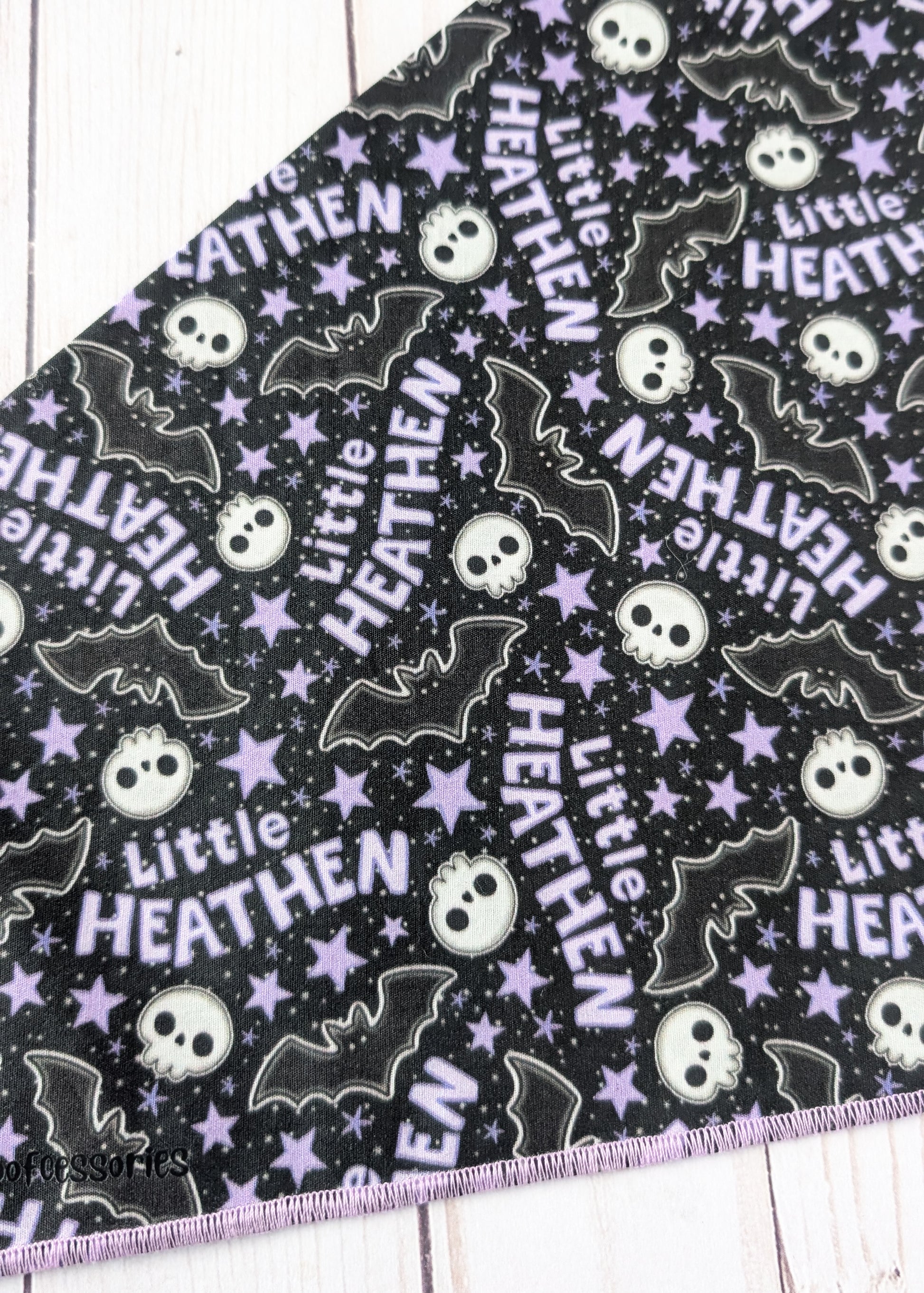 A close-up of a black fabric with a repeating Halloween pattern featuring purple stars, white skulls, black bats, and the text 'Little HEATHEN' in white and purple, laid flat on a white wood surface.