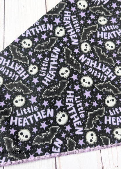 A close-up of a black fabric with a repeating Halloween pattern featuring purple stars, white skulls, black bats, and the text 'Little HEATHEN' in white and purple, laid flat on a white wood surface.