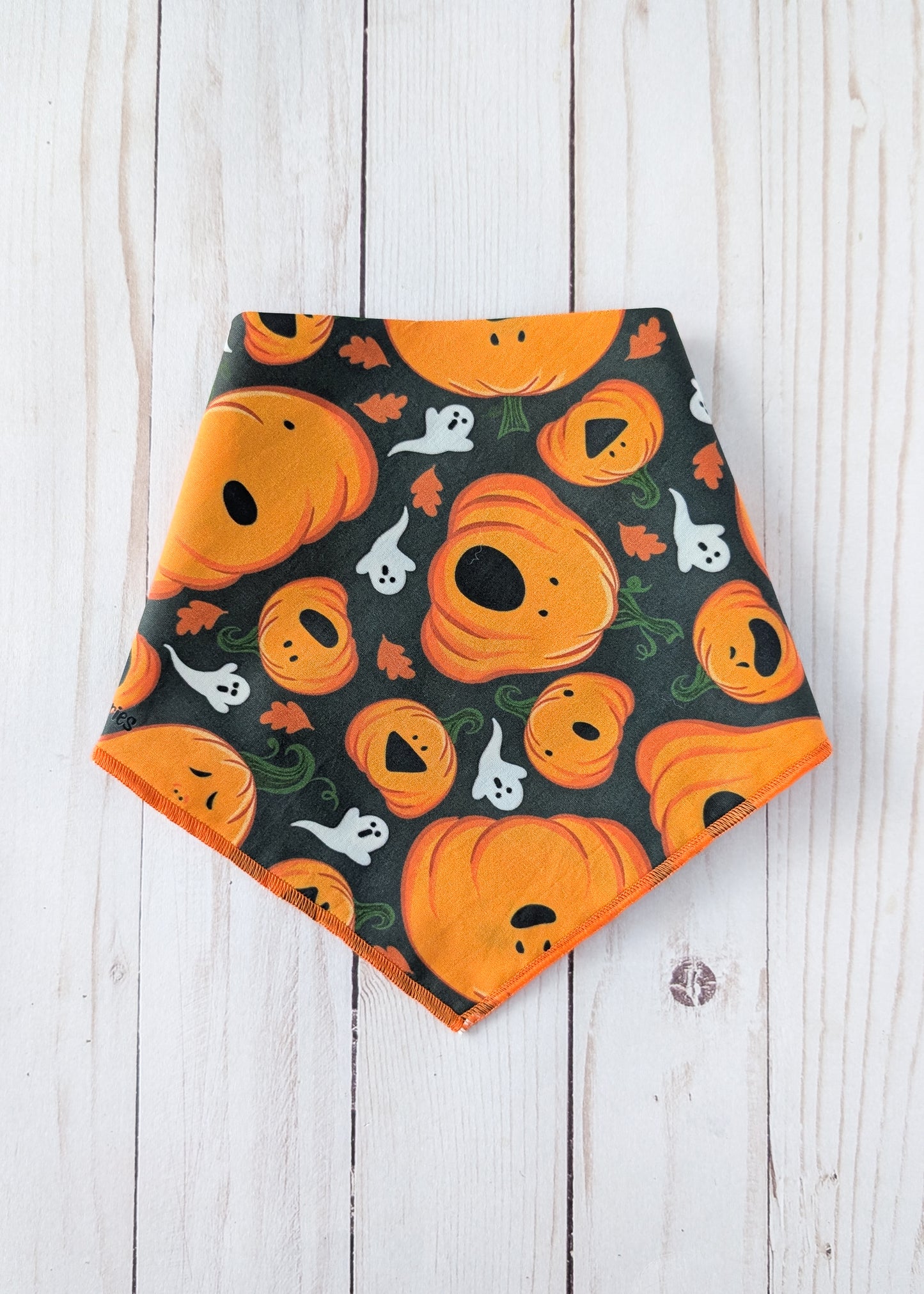 A triangular pet bandana with an orange and black Halloween pattern featuring pumpkins, ghosts, and leaves, laid flat on a white wood plank background.