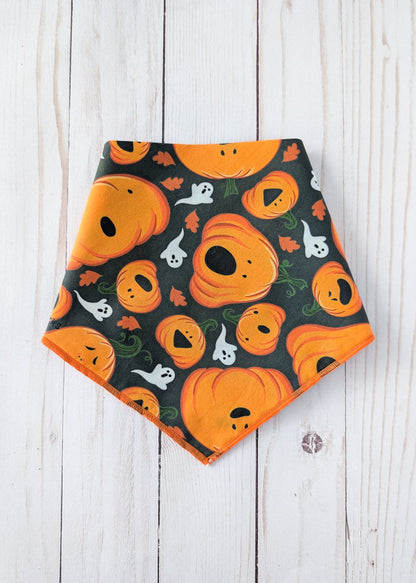 A triangular pet bandana with an orange and black Halloween pattern featuring pumpkins, ghosts, and leaves, laid flat on a white wood plank background.