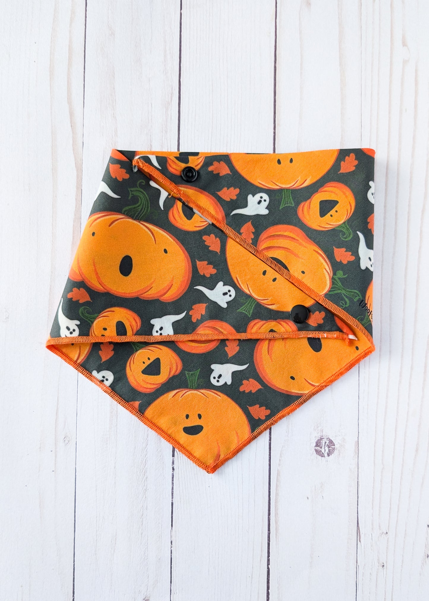 A triangular pet bandana with an orange and black Halloween pattern featuring pumpkins, ghosts, and leaves, laid flat on a white wood plank background.