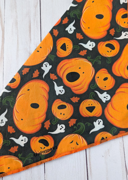 A close up of a triangular pet bandana with an orange and black Halloween pattern featuring pumpkins, ghosts, and leaves, laid flat on a white wood plank background.