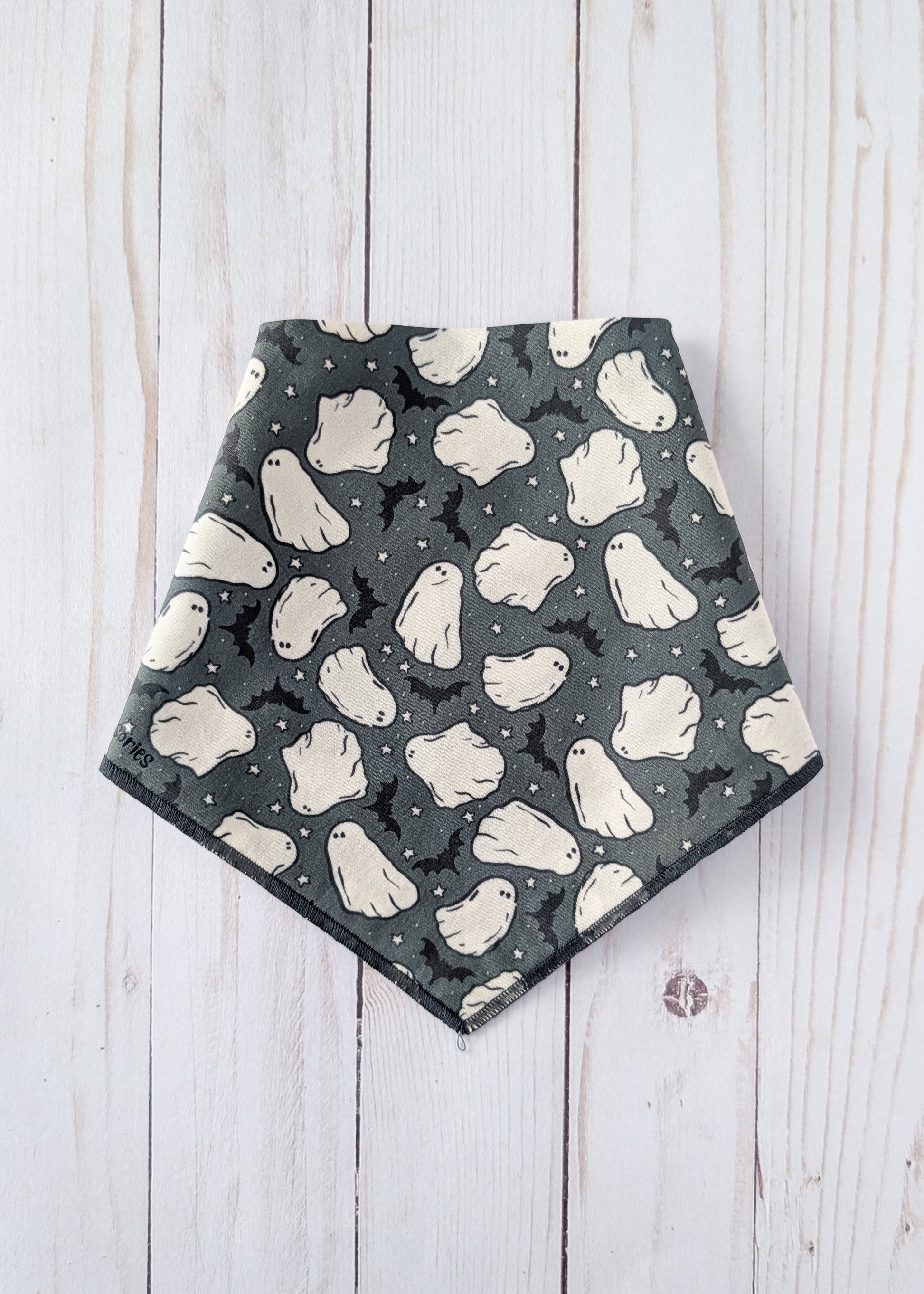 A triangular dog bandana with a Halloween ghost pattern, laid flat on a white wooden plank background.