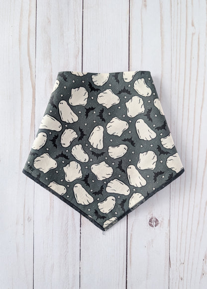 A triangular dog bandana with a Halloween ghost pattern, laid flat on a white wooden plank background.