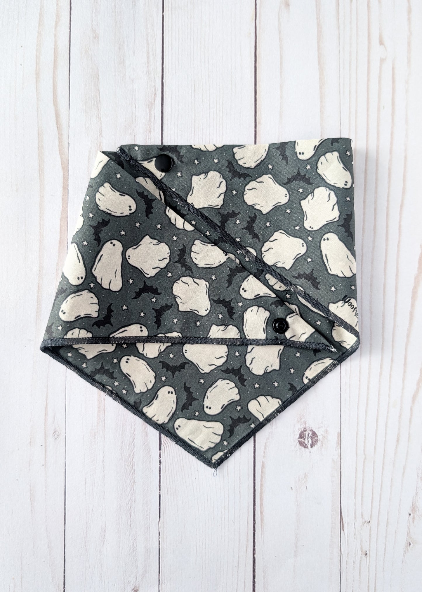 A triangular dog bandana with a Halloween ghost and bat pattern, laid flat on a white wood plank background.