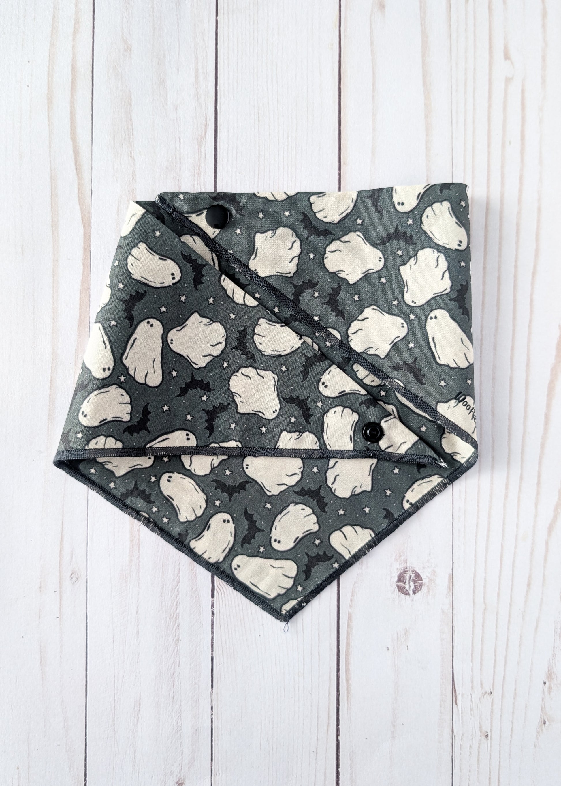 A triangular dog bandana with a Halloween ghost and bat pattern, laid flat on a white wood plank background.