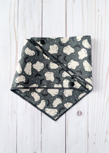 A triangular dog bandana with a Halloween ghost and bat pattern, laid flat on a white wood plank background.
