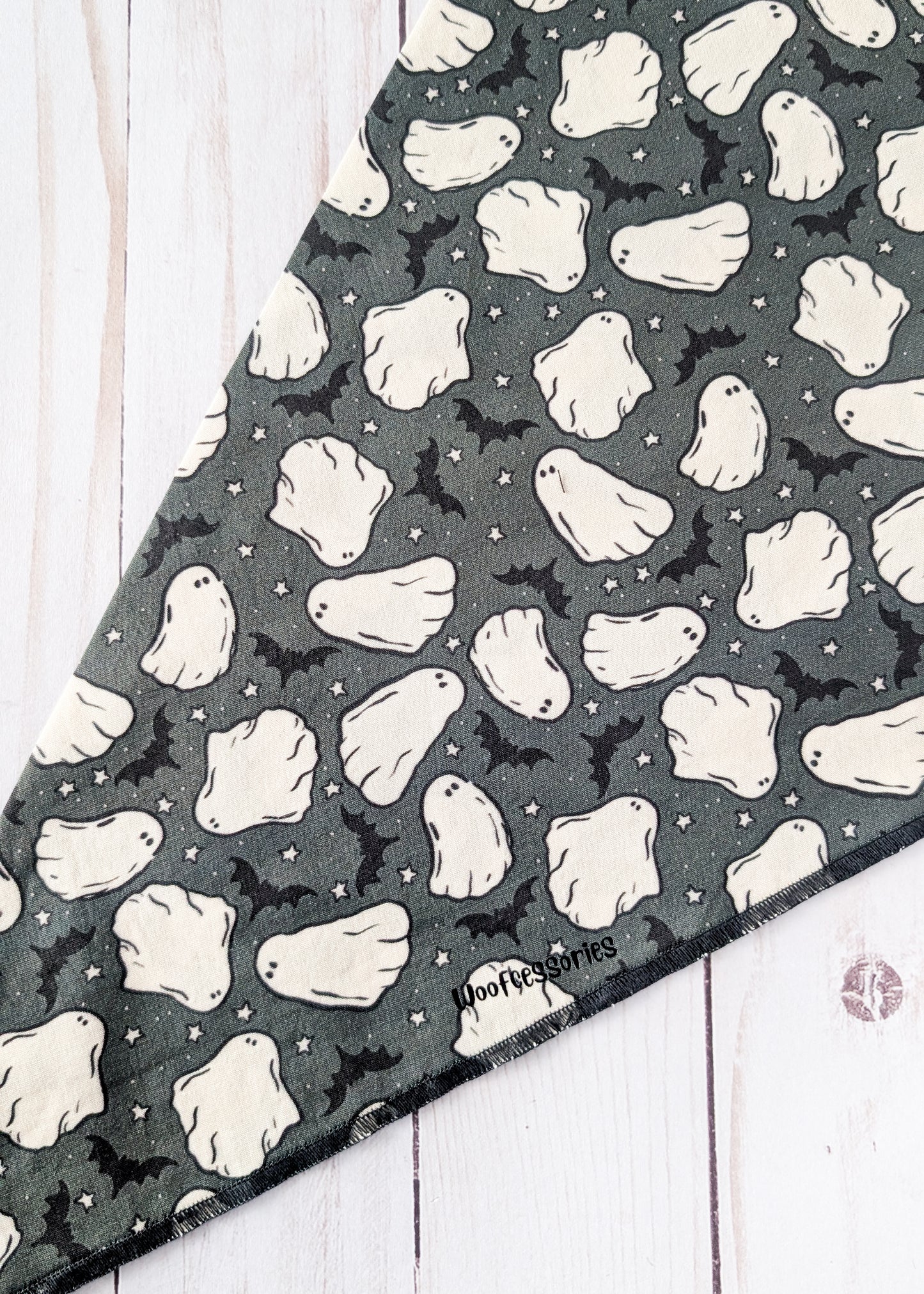 A triangular bandana featuring a Halloween pattern of white ghosts and black bats on a dark gray background, laid flat on a white wood plank surface.