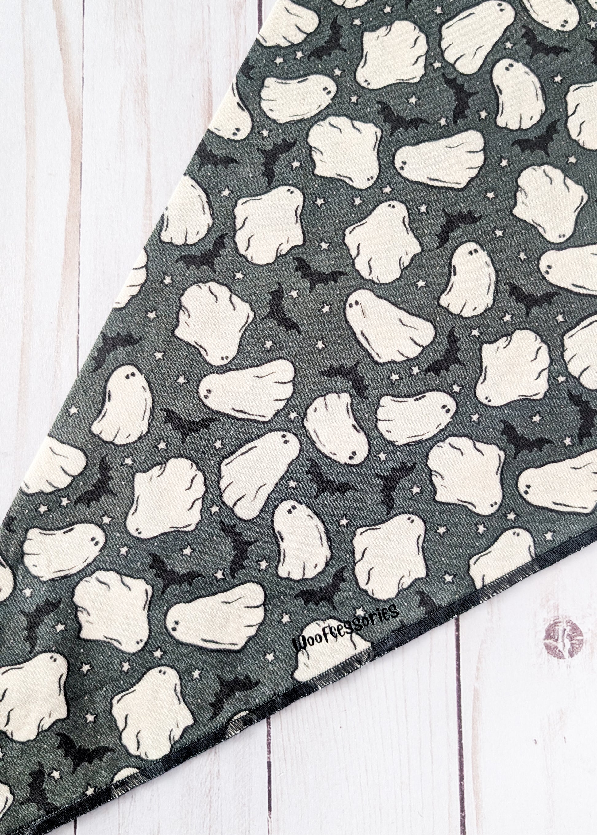 A triangular bandana featuring a Halloween pattern of white ghosts and black bats on a dark gray background, laid flat on a white wood plank surface.