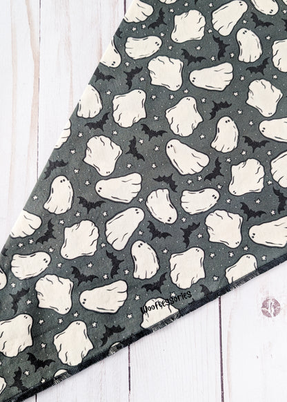 A triangular bandana featuring a Halloween pattern of white ghosts and black bats on a dark gray background, laid flat on a white wood plank surface.