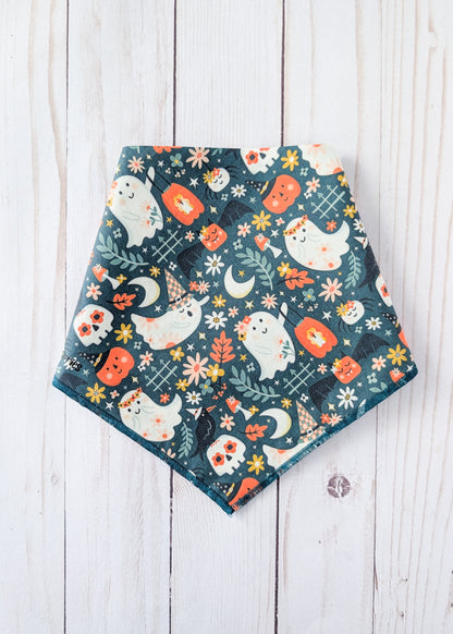 A triangular bandana bib with a Halloween pattern featuring cute white ghosts, orange pumpkins, and cream flowers on a dark teal background, laid flat on a white wooden plank surface.