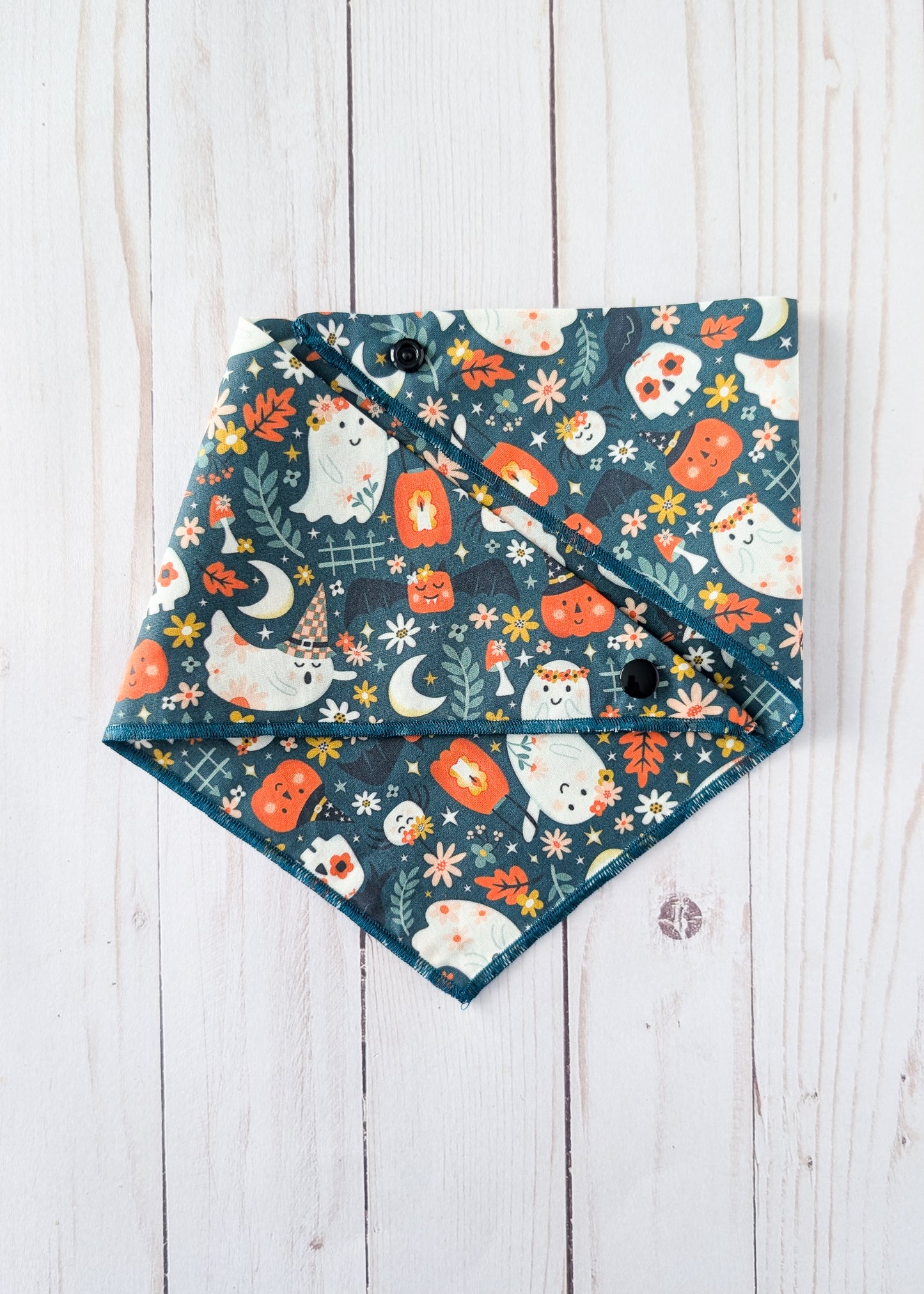 A triangular bandana bib with a Halloween pattern featuring cute white ghosts, orange pumpkins, and cream flowers on a dark teal background, laid flat on a white wooden plank surface.