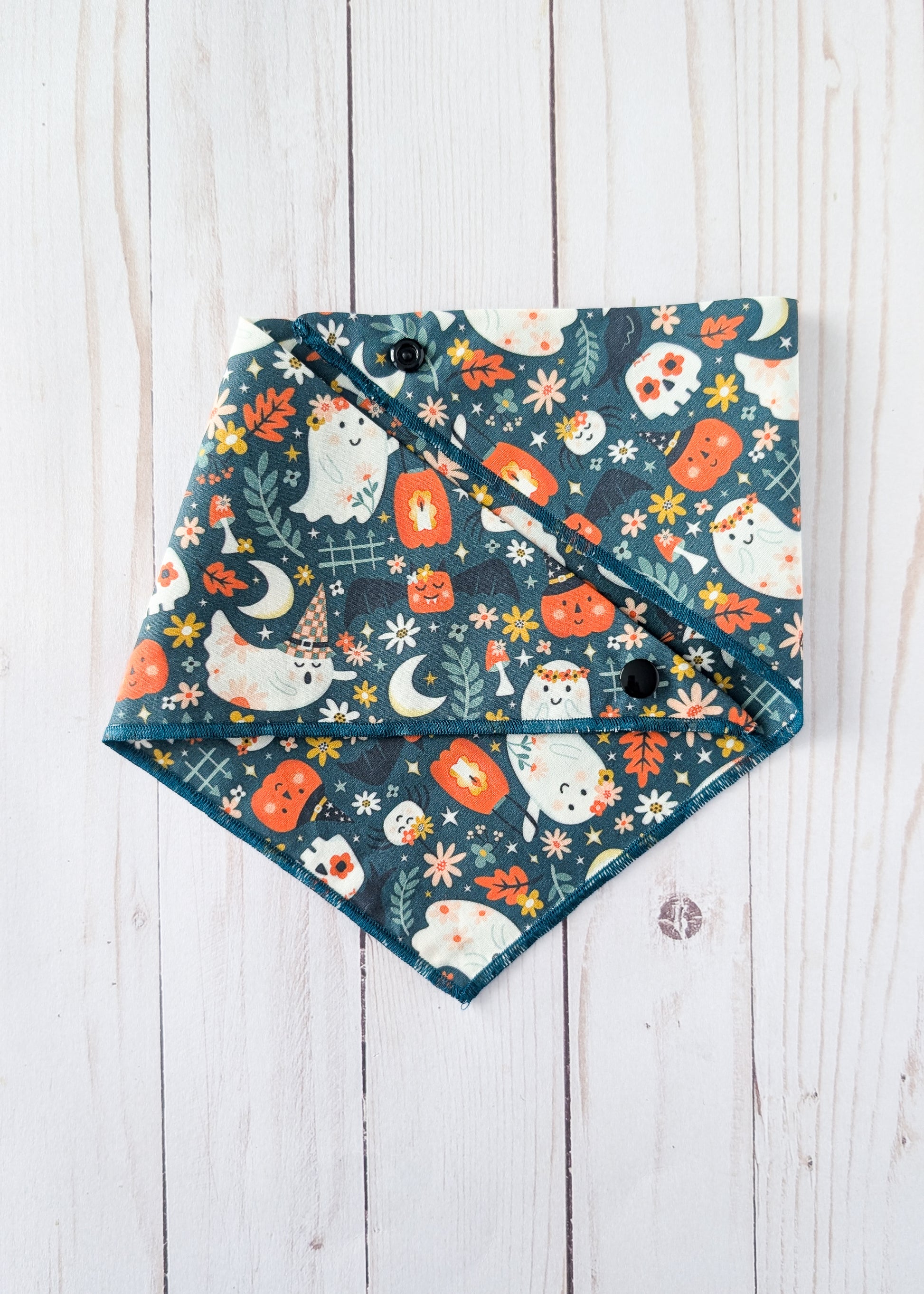 A triangular bandana bib with a Halloween pattern featuring cute white ghosts, orange pumpkins, and cream flowers on a dark teal background, laid flat on a white wooden plank surface.