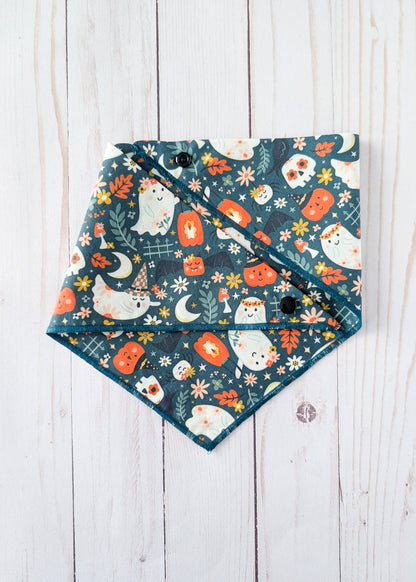 A triangular bandana bib with a Halloween pattern featuring cute white ghosts, orange pumpkins, and cream flowers on a dark teal background, laid flat on a white wooden plank surface.