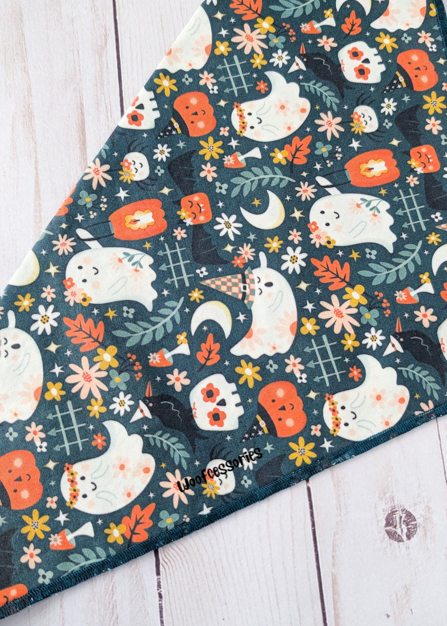 A triangular bandana with a Halloween-themed print of ghosts, pumpkins, flowers, and leaves on a dark teal background, laid flat on a white wooden surface.